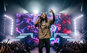 A rapper performs on stage at a concert, with a large screen displaying abstract art behind him and a cheering crowd in front.
