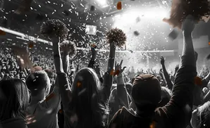 People in a crowd cheer and throw confetti, with some holding pom-poms.