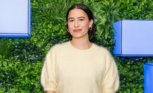 Woman in a cream sweater smiles in front of a green leafy backdrop with blue accents.