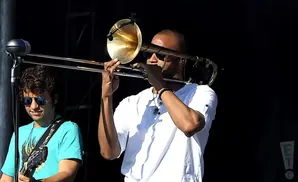 A trombonist plays under bright lights, with a guitarist to his left.