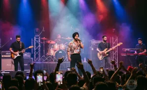 A band performs on stage with colorful lights, a singer in a patterned shirt takes center stage while other members play instruments.