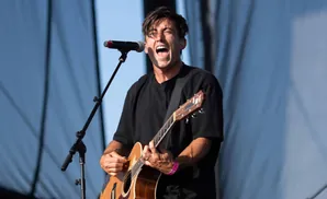 A man with dark hair sings into a microphone while playing an acoustic guitar on a stage.