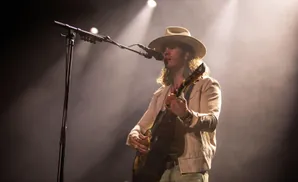 A man in a cowboy hat plays guitar on stage with spotlights shining on him.