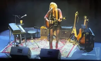 Singer plays acoustic guitar on stage with other guitars and a keyboard.