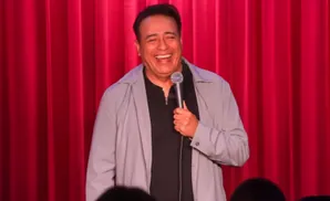 A man in a grey jacket and black shirt laughs into a microphone on a red curtain background.
