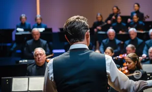 Orchestra conductor leading musicians, with a focus on the conductor's back and the musicians' faces and instruments.