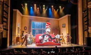 The Cat in the Hat drives a car on stage during a performance, with actors and an orchestra present.