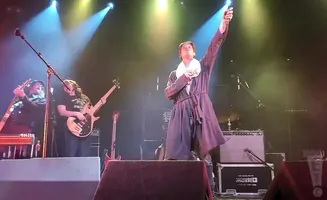 A lead singer in a robe raises a microphone and bell onstage while musicians play guitars.