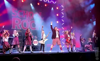 Students in plaid school uniforms rock out on stage at a School of Rock performance.