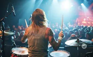 A drummer plays on stage with a crowd in the background under bright stage lights.
