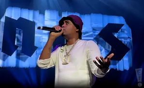 Rapper Nas wearing a white long-sleeve shirt, burgundy bucket hat, and large chain performs on stage.