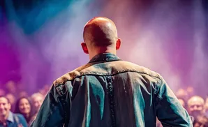 A bald man in a denim shirt faces away from the camera on a stage, with a cheering crowd and purple stage lights behind him.