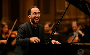 A male pianist with glasses laughs joyfully while playing a grand piano with an orchestra behind him.