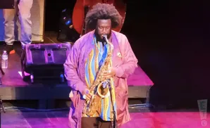 Musician playing a saxophone on stage, wearing a purple jacket and colorful shirt.