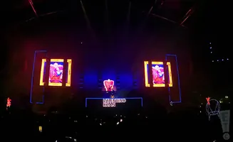 A DJ performs on a stage with large screens displaying graphics, under bright stage lights.