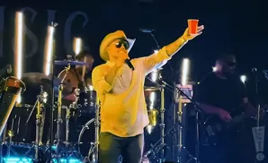 A man in a cowboy hat sings into a microphone, holding up a red cup.