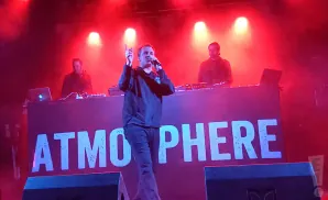 Rapper Slug from Atmosphere performs on stage with a DJ and another band member under red lights.