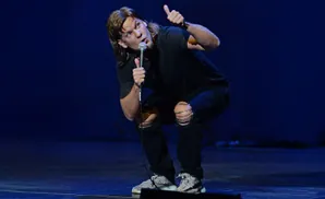 Comedian Chris D'Elia squats on stage with a microphone and gives a thumbs up.
