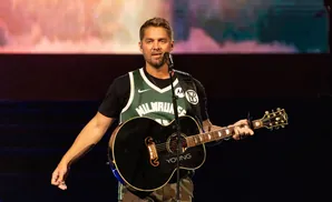 A man in a green basketball jersey and camouflage pants plays an acoustic guitar on stage.