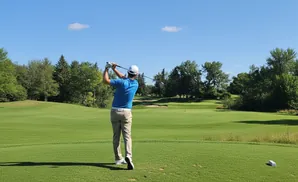 Golfer in blue shirt swings a club on a golf course with green grass and trees.