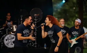 A band performs on stage, with two singers in the foreground and a guitarist and drummer in the background.