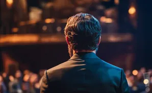 Man in a dark suit seen from behind, facing a blurred audience under stage lights.