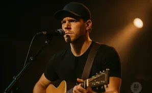 A man in a black cap and t-shirt plays an acoustic guitar and sings into a microphone.