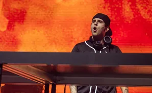 A male DJ wearing a black baseball cap and jersey with headphones around his neck performs on a stage with an orange, blurred background.