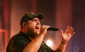 A man wearing a camo trucker hat sings into a microphone with his eyes closed and one hand raised.