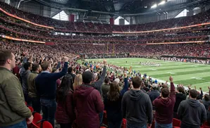 Fans cheer at a football game in a stadium with a green field and red seats.
