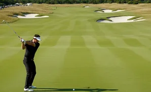 Golfer in black mid-swing on a bright green golf course with sand traps and spectators in the background.