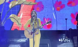 A woman in a yellow dress plays an acoustic guitar and sings into a microphone on stage.