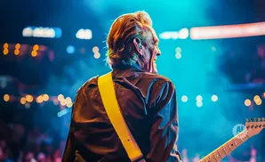 Musician with blonde hair and yellow guitar strap playing a guitar on stage with bright lights.