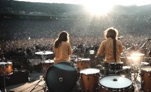 Two drummers perform on stage at a large outdoor concert, silhouetted against the setting sun.