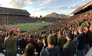 Spectators cheer at a football game in a packed stadium with a mountain range in the background.