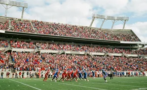 Football players in red and blue uniforms on a field, with a large crowd in the stands of a stadium under a cloudy sky.