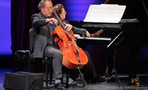 A man plays a cello next to a woman playing a piano, on a stage with purple and blue lighting.