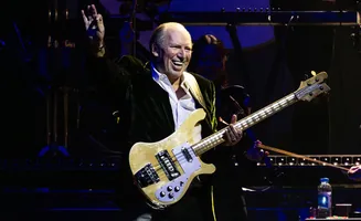 Musician playing a bass guitar on stage, waving to the audience.