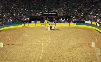 A rodeo rider on a horse is near a barrel in a dirt arena, with spectators in the background.