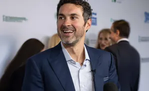 Man in a blue suit smiles at a microphone, with blurred people in the background.