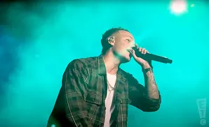 A rapper wearing a plaid shirt and gold chain performs into a microphone on a stage lit with green lights.