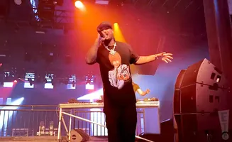 A rapper with a large pendant necklace performs on stage, illuminated by vibrant stage lights.