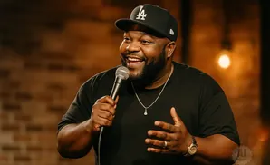 A Black man in a LA Dodgers baseball cap and black t-shirt smiles while holding a microphone and gesturing with his hands.