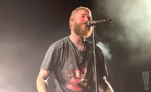 Post Malone sings into a microphone on a smoky stage, wearing a black t-shirt with a graphic print.