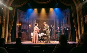 A stage play scene with actors in Victorian attire performing on a set depicting a richly decorated room.