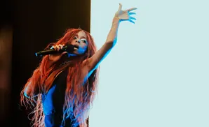Singer with long red hair on stage, holding a microphone and reaching up with her other hand.