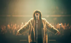 A man in a leopard print coat sings into a microphone on stage with a blurred audience behind him.