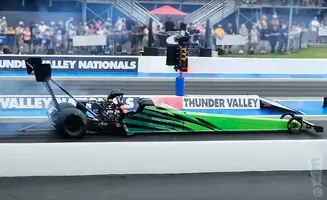 A green and black dragster races on a track with a crowd in the background.