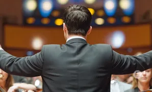 A conductor in a black suit faces away from the camera, arms outstretched, leading an orchestra.