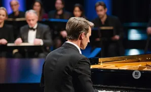 A pianist in a tuxedo plays a grand piano with an orchestra behind him.